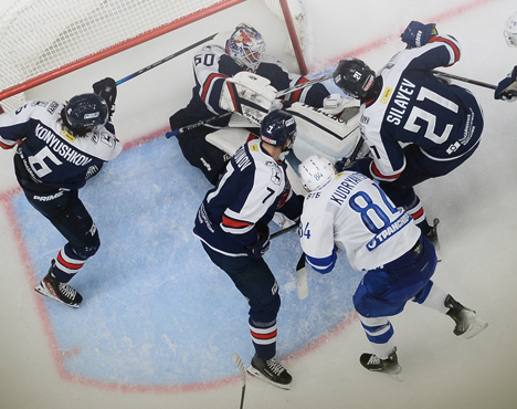 Хоккейный матч: пять игроков в бело-синих формах и вратарь в черной форме борются за шайбу у ворот. Вратарь защищает ворота, игроки противоборствующих команд активно участвуют в борьбе. Сцена динамичная и напряженная